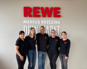 Fünf Mitarbeiter:innen stehen vor dem Firmenlogo des Rewe Markus Brzezina.