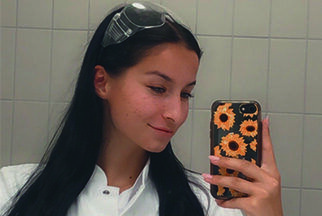 Eine junge Frau mit dunklen Haaren steht in einem Laborkittel fotografiert ihr Spiegelbild mit einem Handy.