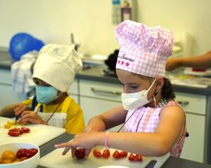 Ein kliener junge und ein kleines Mädchen mit Kochmützen, Mundschutz und Schürzen schneiden in einer Lehrküche Tomaten.