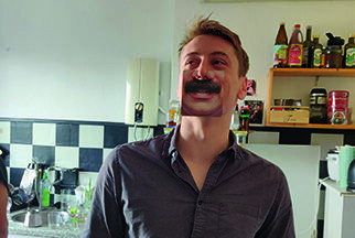 Stammzellspender Klemens steht mit einem aufgeklebten Schnurrbart in einer Küche.