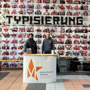 Stand der Stefan Morsch Stiftung mit zwei Mitarbeitern vor der Fanwand des 1.FC Kaiserslautern