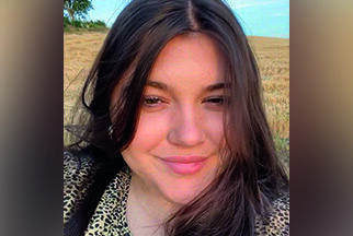 Eine junge Frau mit langen braunen Haaren steht vor einem Feld und lächelt.