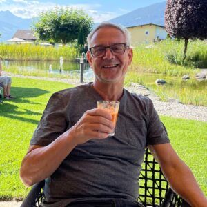Ein grauhaariger Mann sitzt lächelnd in sommerlicher Kleidung auf einer Terrasse mit einem Getränk in der Hand