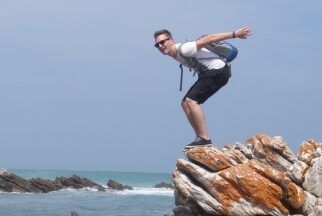 Ein Mann mit Rucksack und Sonnenbrille deutet einen Sprung von einem Fels an.