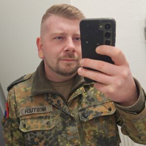 Ein Mann in Bundeswehrkleidung steht vor einem Spiegel und fotografiert sich mit einem Smartphone.